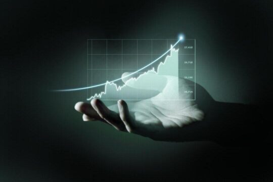 Uma mão aberta em um fundo escuro projeta um holograma com um gráfico de crescimento financeiro. A linha do gráfico tem um formato ascendente, com um ponto luminoso no topo, indicando uma tendência positiva.