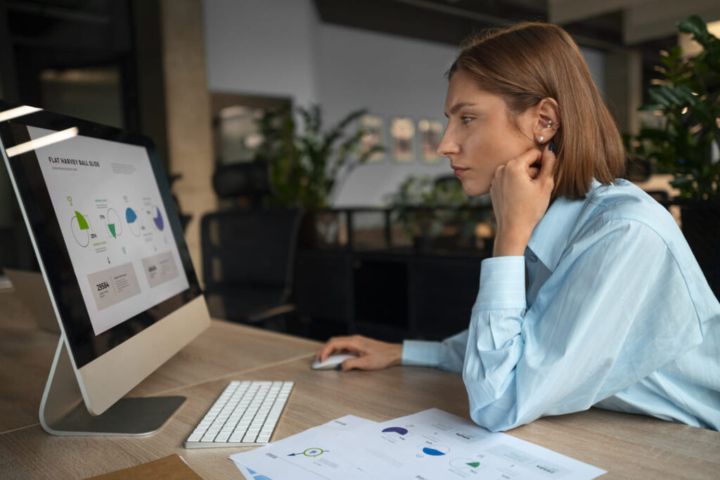A imagem mostra uma mulher jovem, de cabelos castanhos e lisos, usando uma camisa azul-clara, sentada em uma mesa de escritório. Ela observa atentamente um monitor de computador que exibe gráficos coloridos em formato de pizza e barras. Sobre a mesa, há também folhas impressas com os mesmos gráficos. O ambiente é moderno, com iluminação suave e plantas ao fundo, sugerindo um espaço de trabalho organizado e voltado para o uso de ferramentas analíticas e automação de processos.