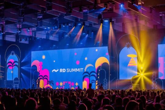 Palco principal iluminado do RD Summit 2024, com telão colorido exibindo o logotipo do evento diante de uma plateia cheia, representando um dos maiores encontros de marketing e inovação da América Latina.