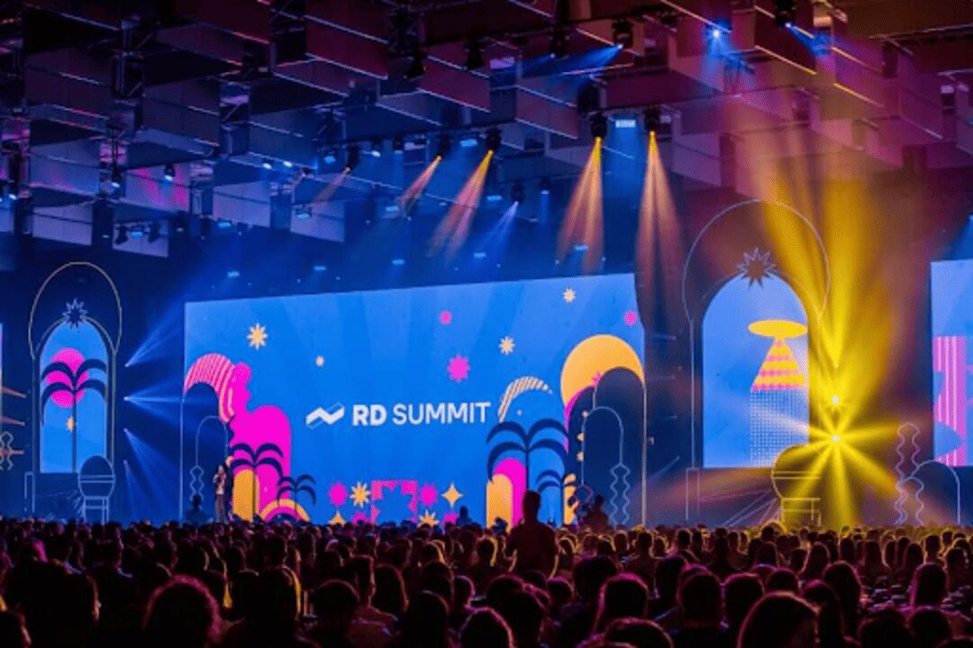 Palco principal iluminado do RD Summit 2024, com telão colorido exibindo o logotipo do evento diante de uma plateia cheia, representando um dos maiores encontros de marketing e inovação da América Latina.