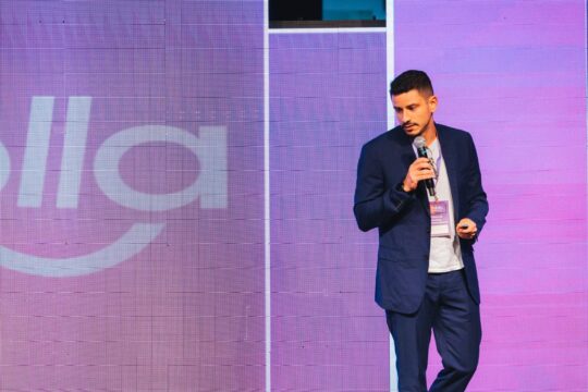 Zé Luiz em palco durante palestra, vestindo terno azul e segurando microfone, em frente a um painel digital com fundo roxo e iluminação de evento corporativo.