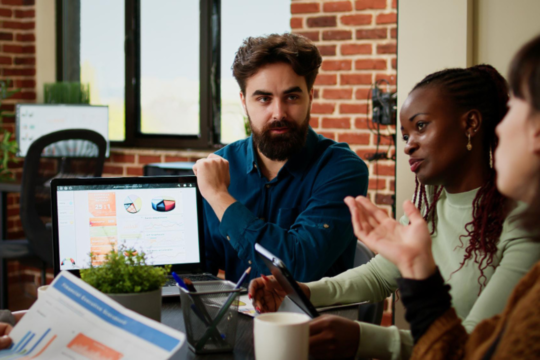 Grupo de profissionais em reunião analisando gráficos e dados de marketing em um laptop, discutindo estratégias digitais em um ambiente de escritório moderno.