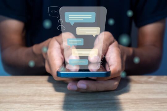 “Mãos segurando um smartphone sobre mesa de madeira; balões de conversa translúcidos em azul e creme flutuam acima da tela, sugerindo troca de mensagens e chatbots — conceito de parceria GMZ.MOKE + Blip.