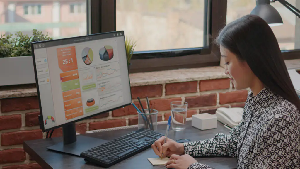 A imagem mostra uma mulher em um escritório com paredes de tijolo aparente. Ela está escrevendo em um bloco de notas enquanto olha para um monitor de computador grande que exibe gráficos de pizza, gráficos de linha e indicadores numéricos relacionados a análises de negócios, como oportunidades, propostas e contratos assinados. A cena é iluminada por luz natural vinda de uma janela ao fundo, e há itens como teclado, copo de água, suporte para canetas e um telefone ao redor da mesa, sugerindo um ambiente de trabalho analítico e organizado.